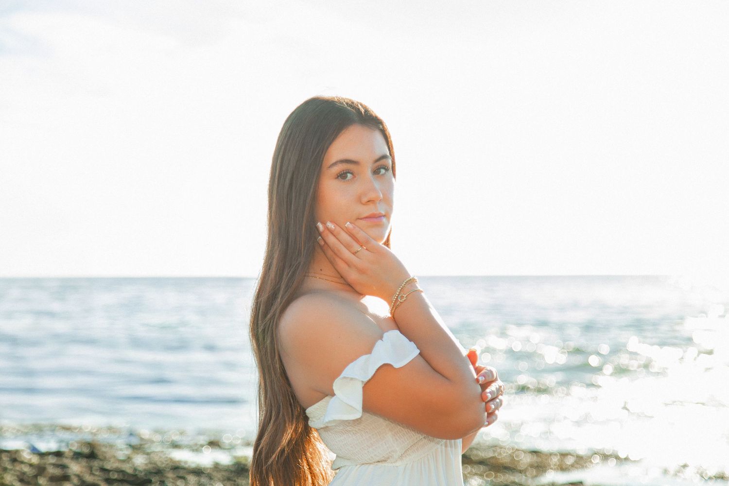 Someone in a white dress poses thoughtfully by the ocean shore during golden hour with sparkling water in background.