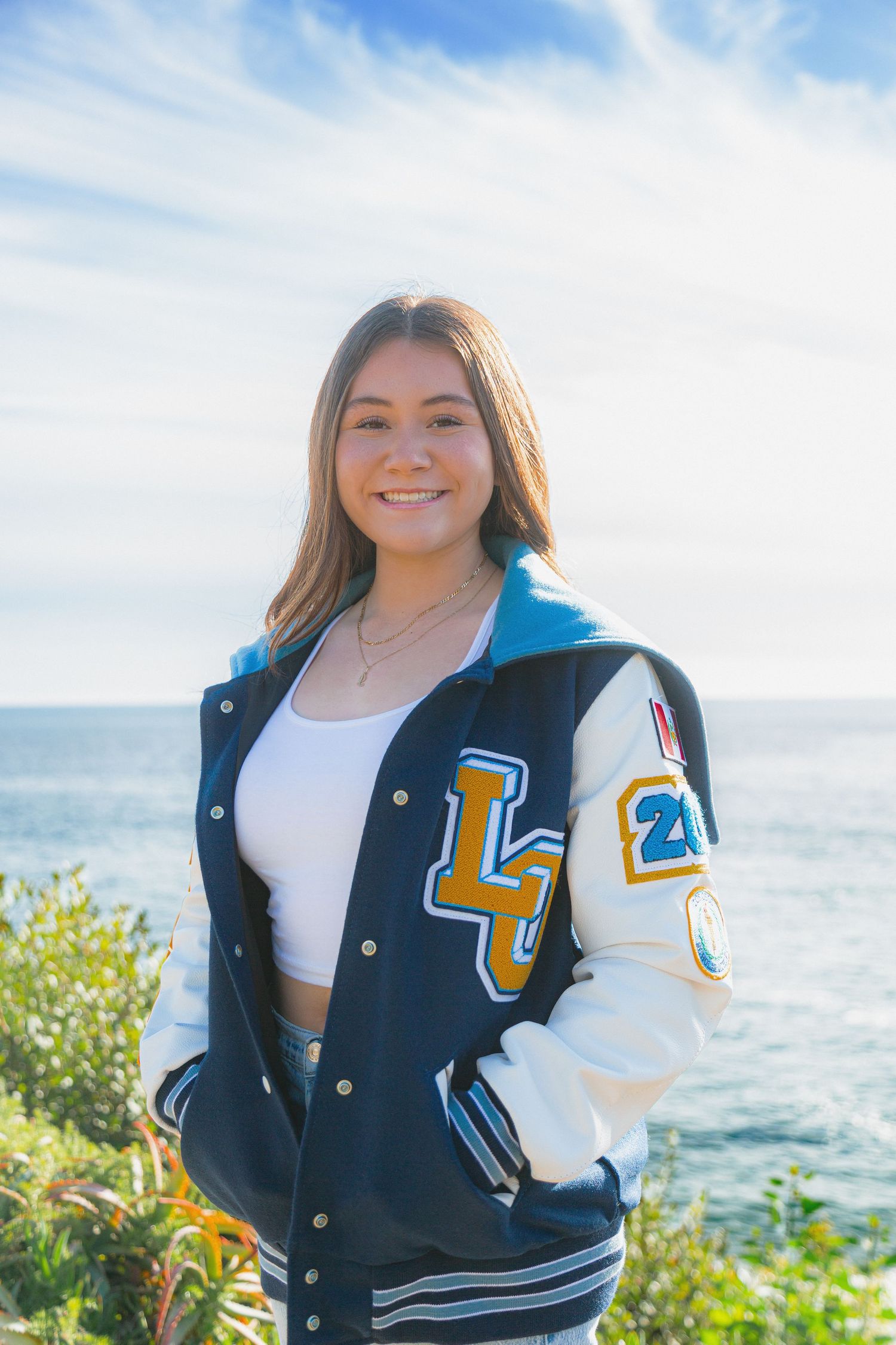 Four photos of someone wearing a navy and white varsity letterman jacket posing outdoors by the ocean on a sunny day.
