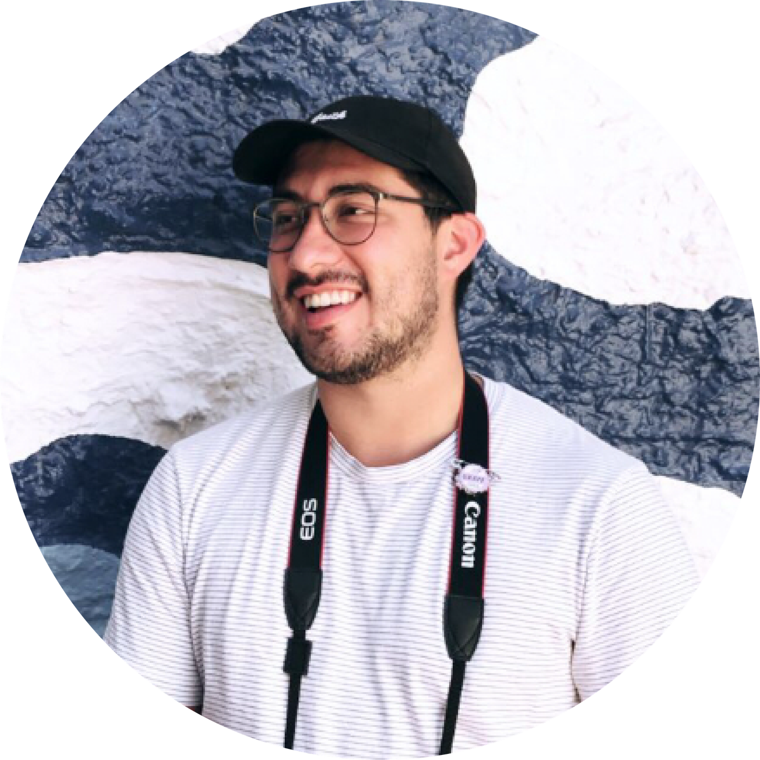 Person wearing glasses and a black cap smiles while wearing a white striped shirt against a blue and white background.