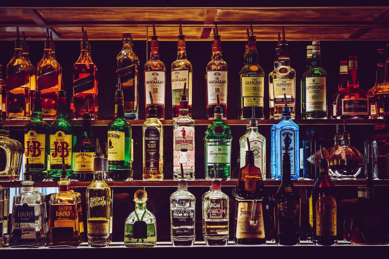 Illuminated liquor bottles line a dark bar shelf with warm ambient lighting creating a cozy atmosphere.