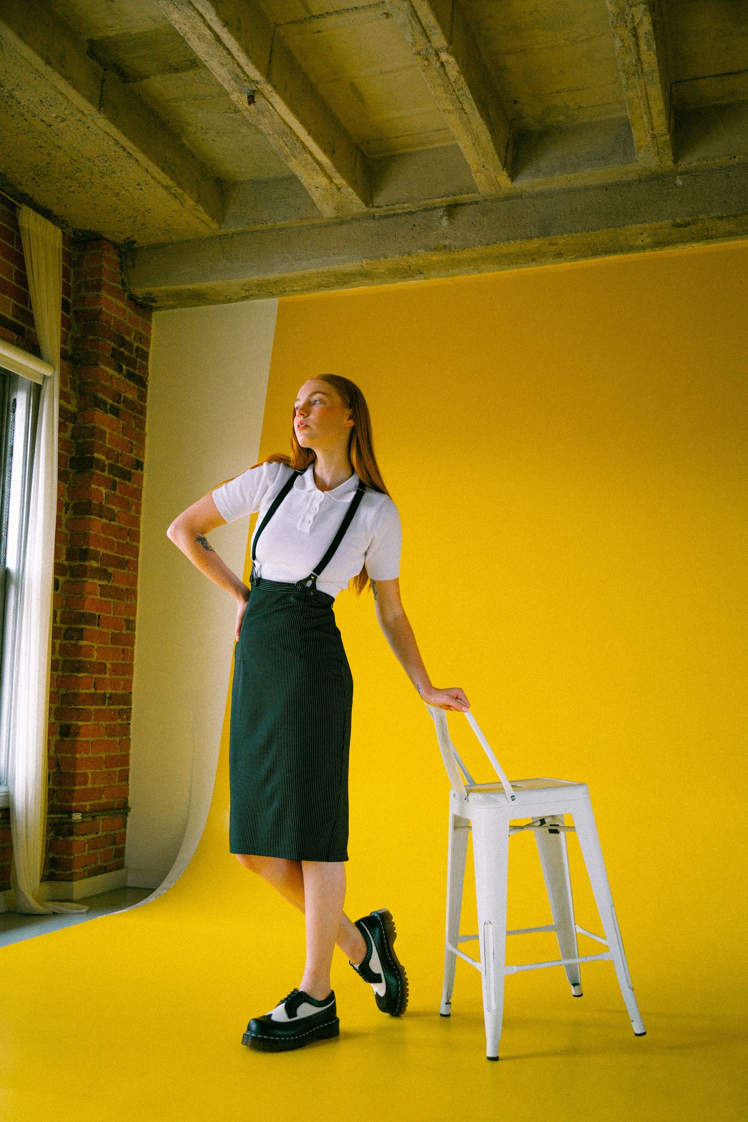 Fashion photo against bright yellow backdrop with white stool, brick wall and wooden beams showing retro school uniform style.