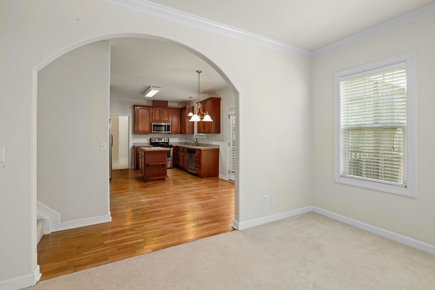 Interior view of an empty home with hardwood floors, white walls, and arched doorway leading to kitchen area.