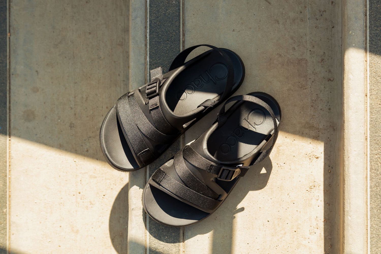A pair of black leather sandals casting shadows on a light surface in natural sunlight.