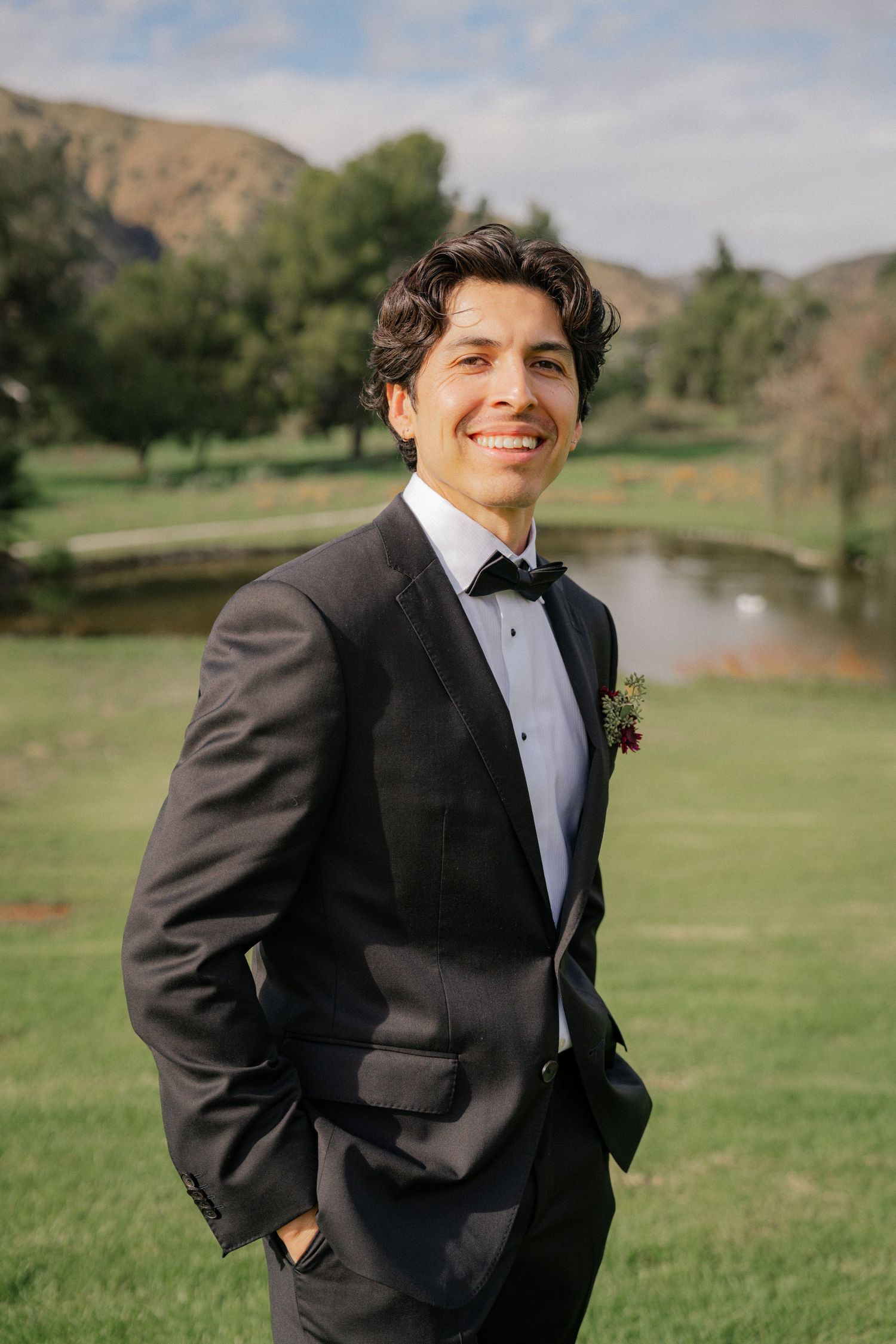 A person in a black tuxedo and bowtie poses outdoors on a golf course with mountains in the background.