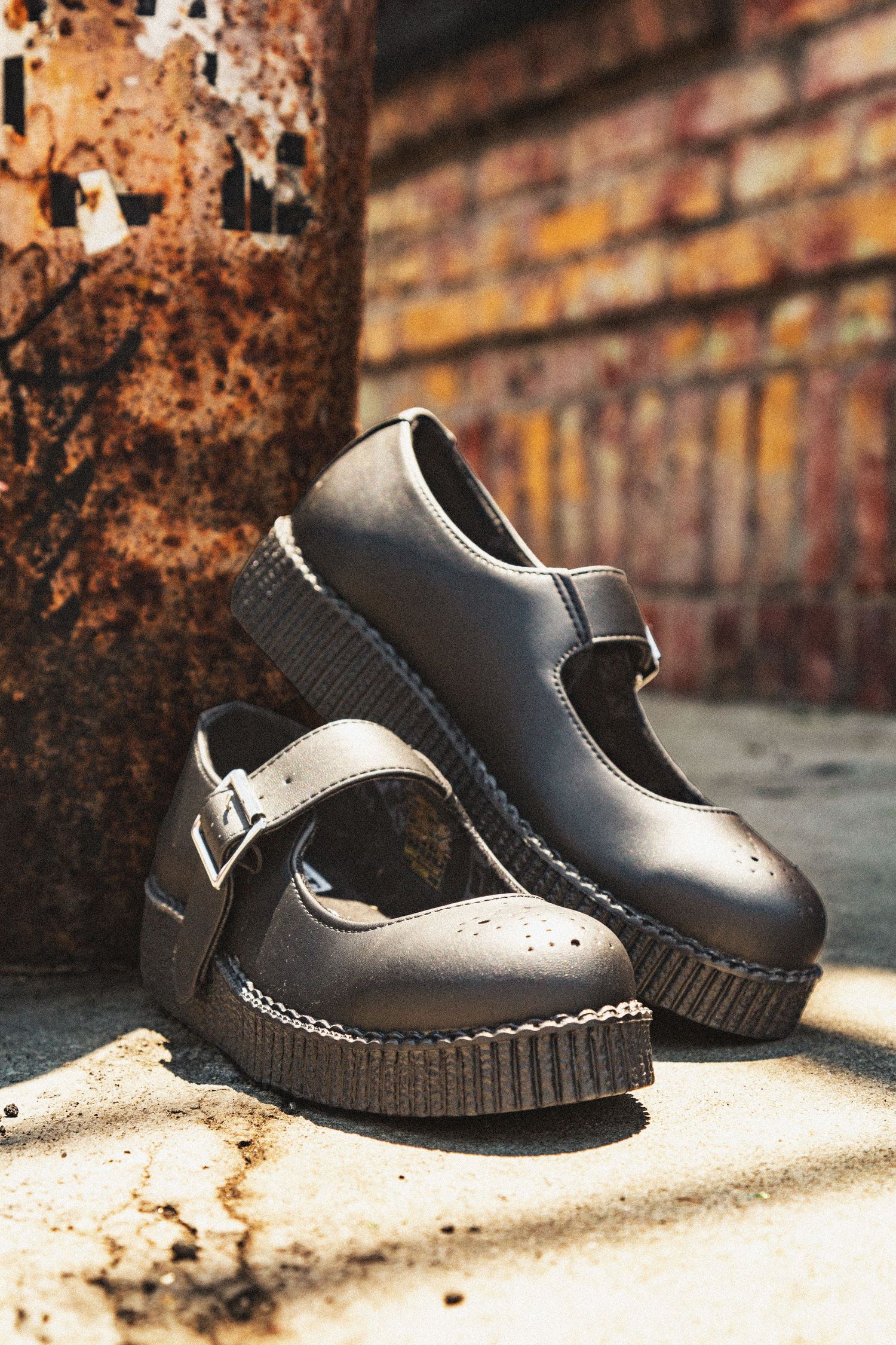 Black leather creeper shoes with white stitching displayed in sequence against a rustic brick wall background.