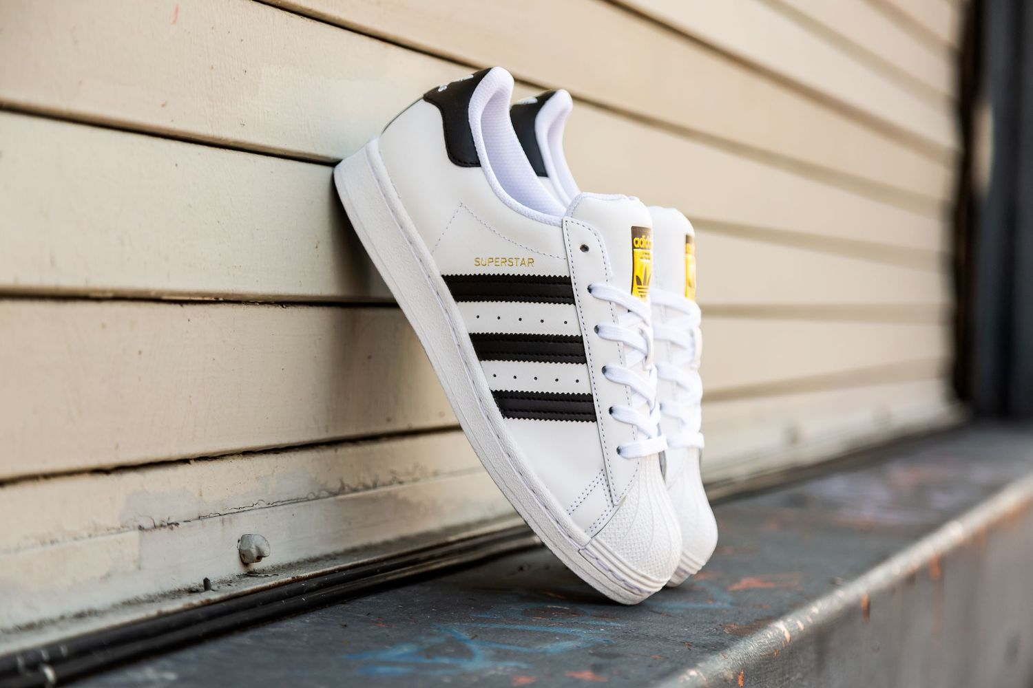 White Adidas Superstar sneaker with black stripes balanced on a curved metal handrail against a beige wall.