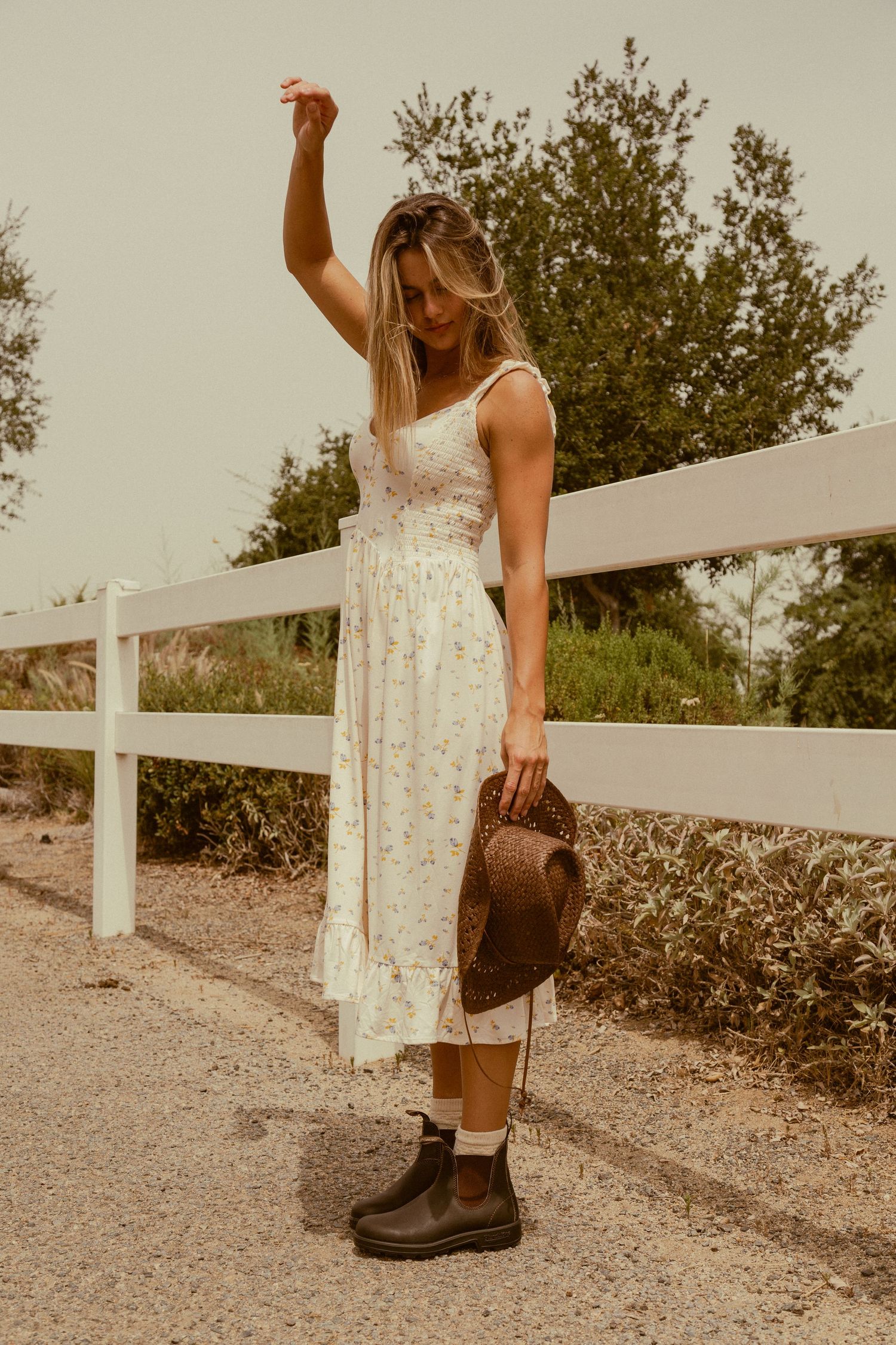 A person in a white lace dress and combat boots stands by a white fence holding a brown hat at sunset.