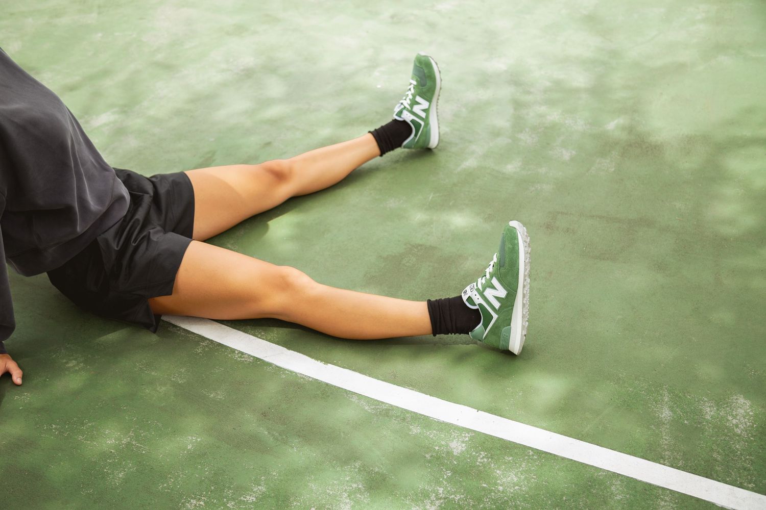 Athletic legs stretched out on a green tennis court surface with white line markings and gray sneakers.