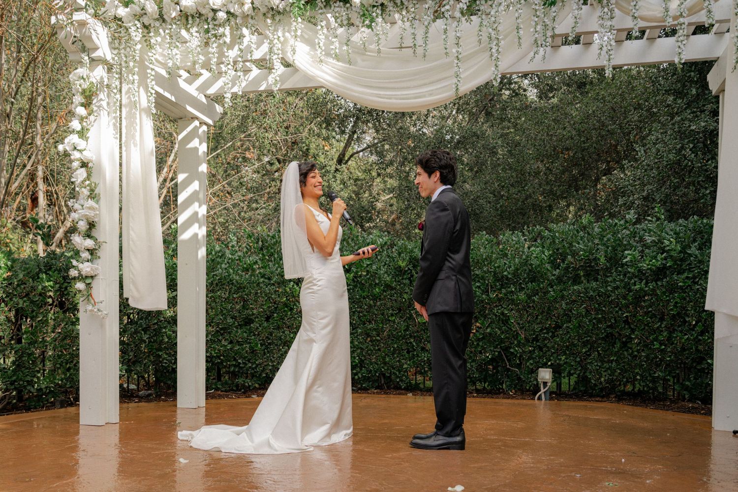 Wedding ceremony taking place in an elegant outdoor gazebo surrounded by lush greenery and white columns.