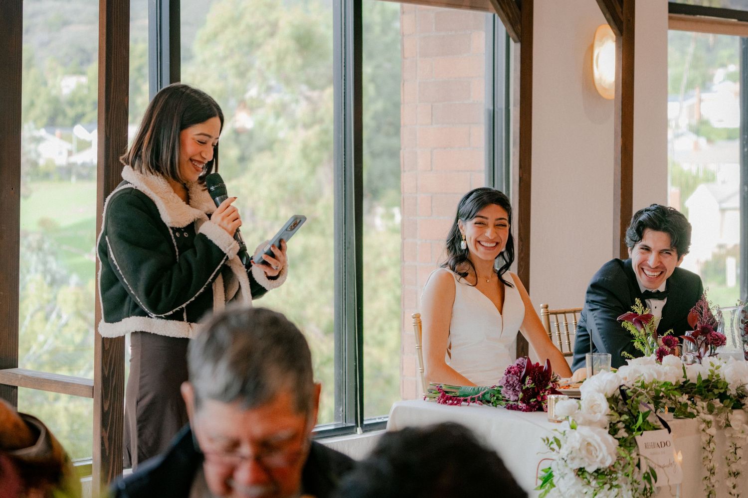 A group of people gathered at a table by large windows during a daytime event with floral arrangements.
