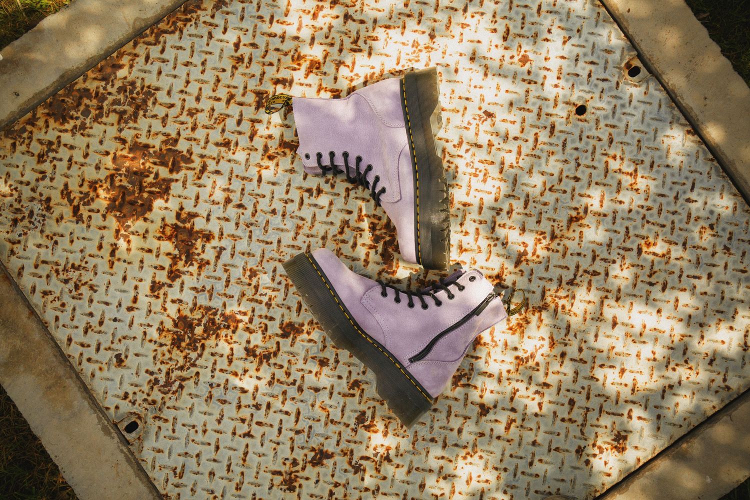 Pair of lavender platform boots with black laces sits on a mosaic tile surface in dappled sunlight.