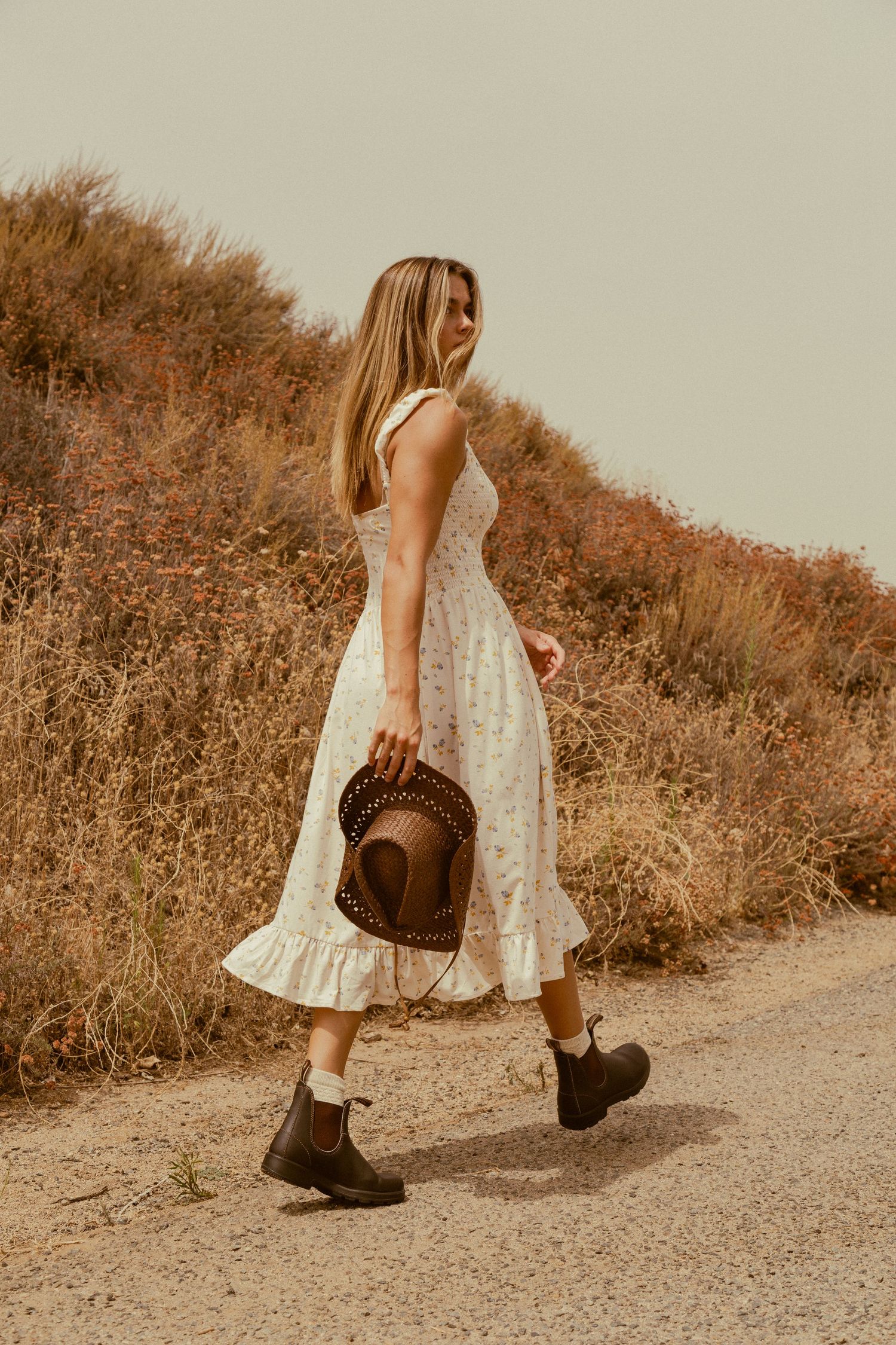 Person in white floral sundress and boots walks on dirt path through golden brown hillside in warm vintage-style photo.