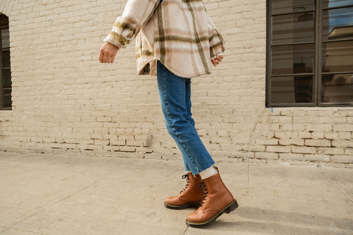 Person wearing brown leather ankle boots and blue jeans stands on white brick sidewalk.