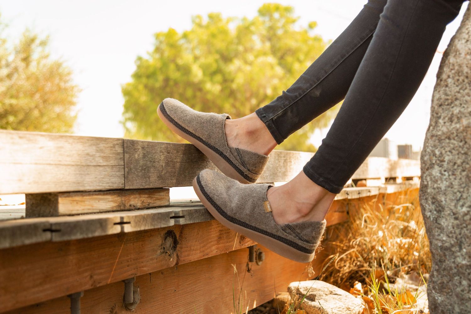Person wearing black jeans and gray suede slip-on shoes sits on wooden deck during golden sunset.