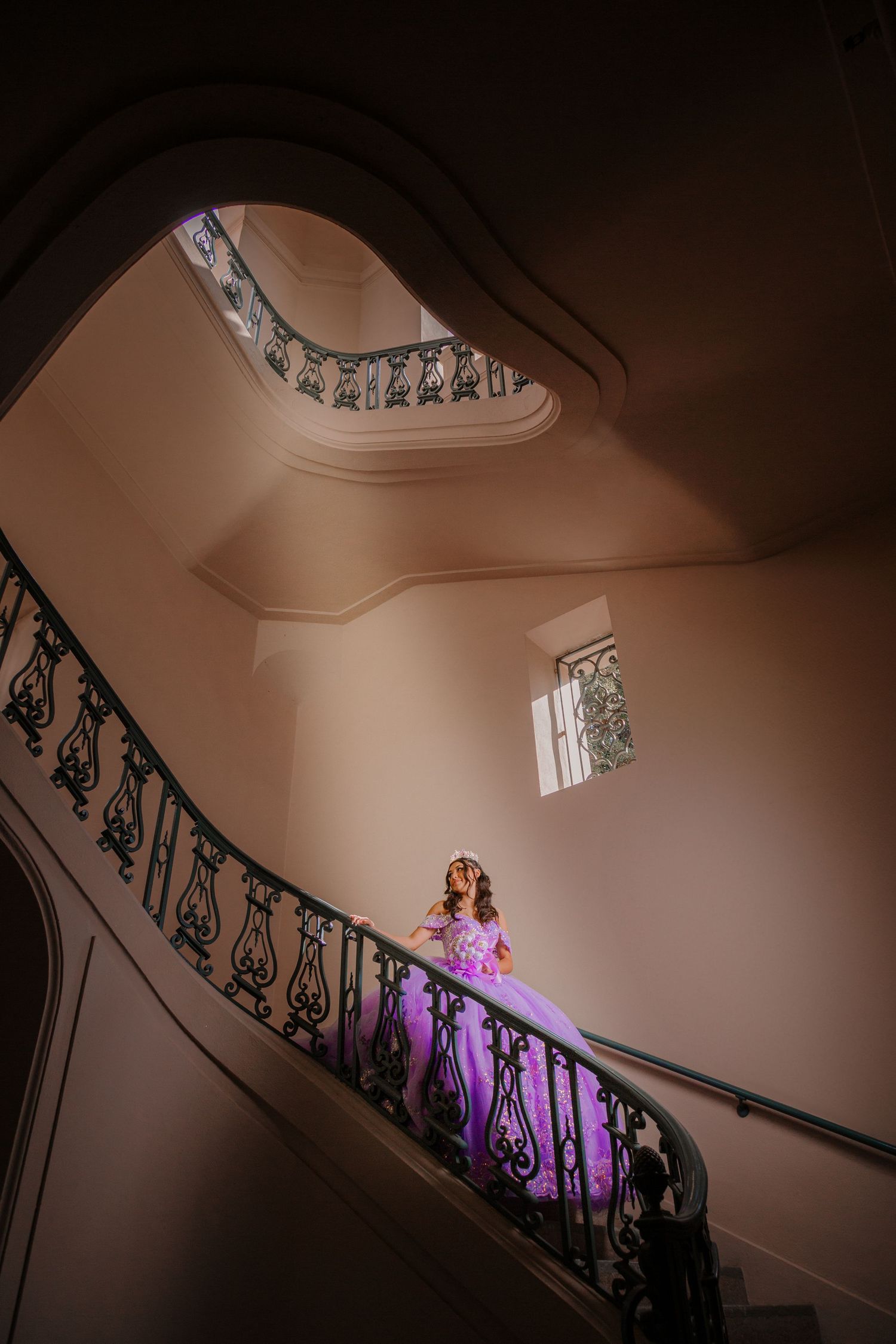 A figure in a flowing purple gown stands on an elegant spiral staircase with ornate black railings in dramatic lighting.