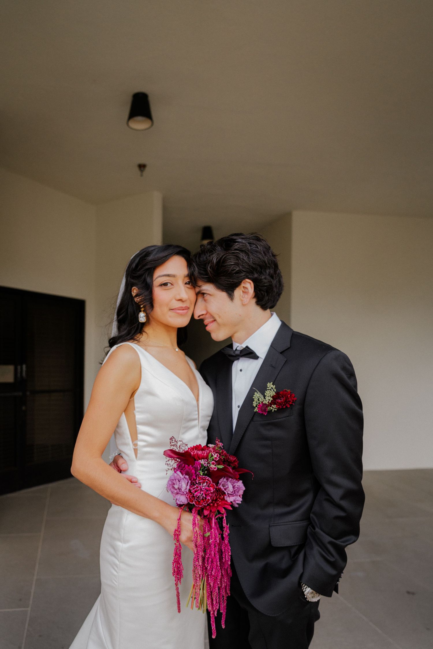 A couple in wedding attire stands together in a hallway, with the bride holding a vibrant magenta bouquet.