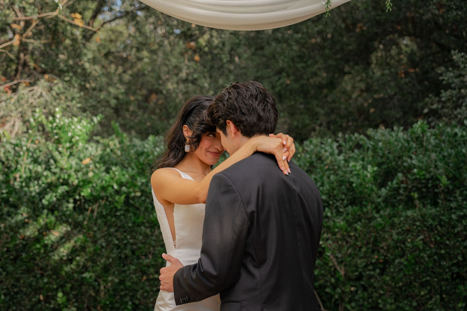 A couple shares an intimate embrace outdoors surrounded by lush green foliage during a wedding photoshoot.