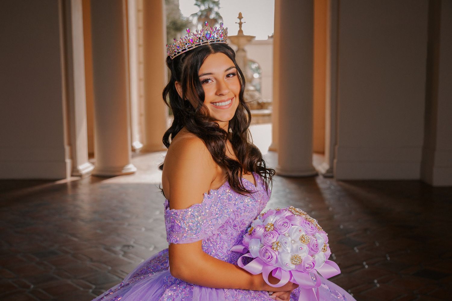 A person in a lavender quinceañera dress poses in an elegant hallway with natural lighting.