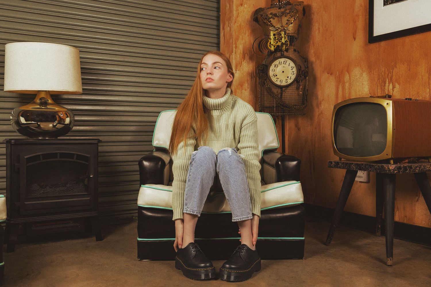 Person relaxes in vintage armchair surrounded by retro decor including old TV and wall clock in wood-paneled room.