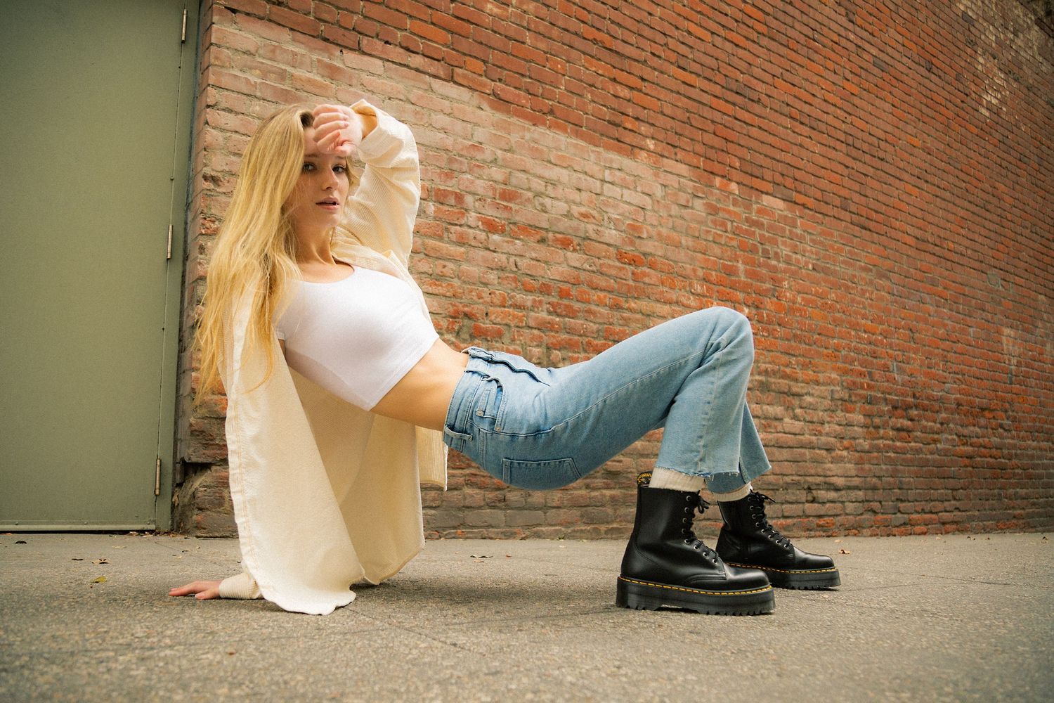 Person in casual attire sits against brick wall with legs outstretched on concrete ground.