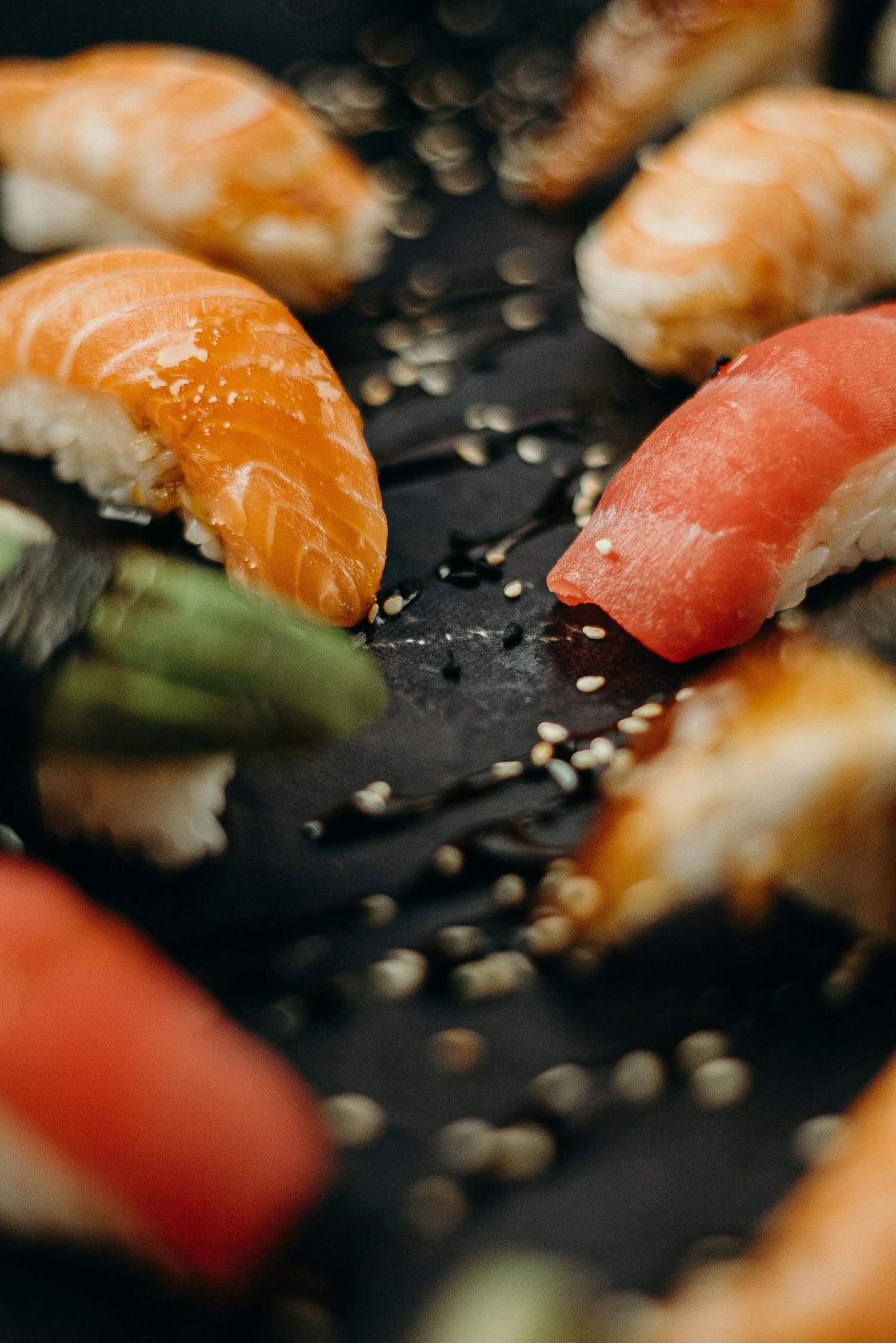 Close-up shot of fresh sushi pieces with salmon and tuna on a dark plate with scattered sesame seeds.