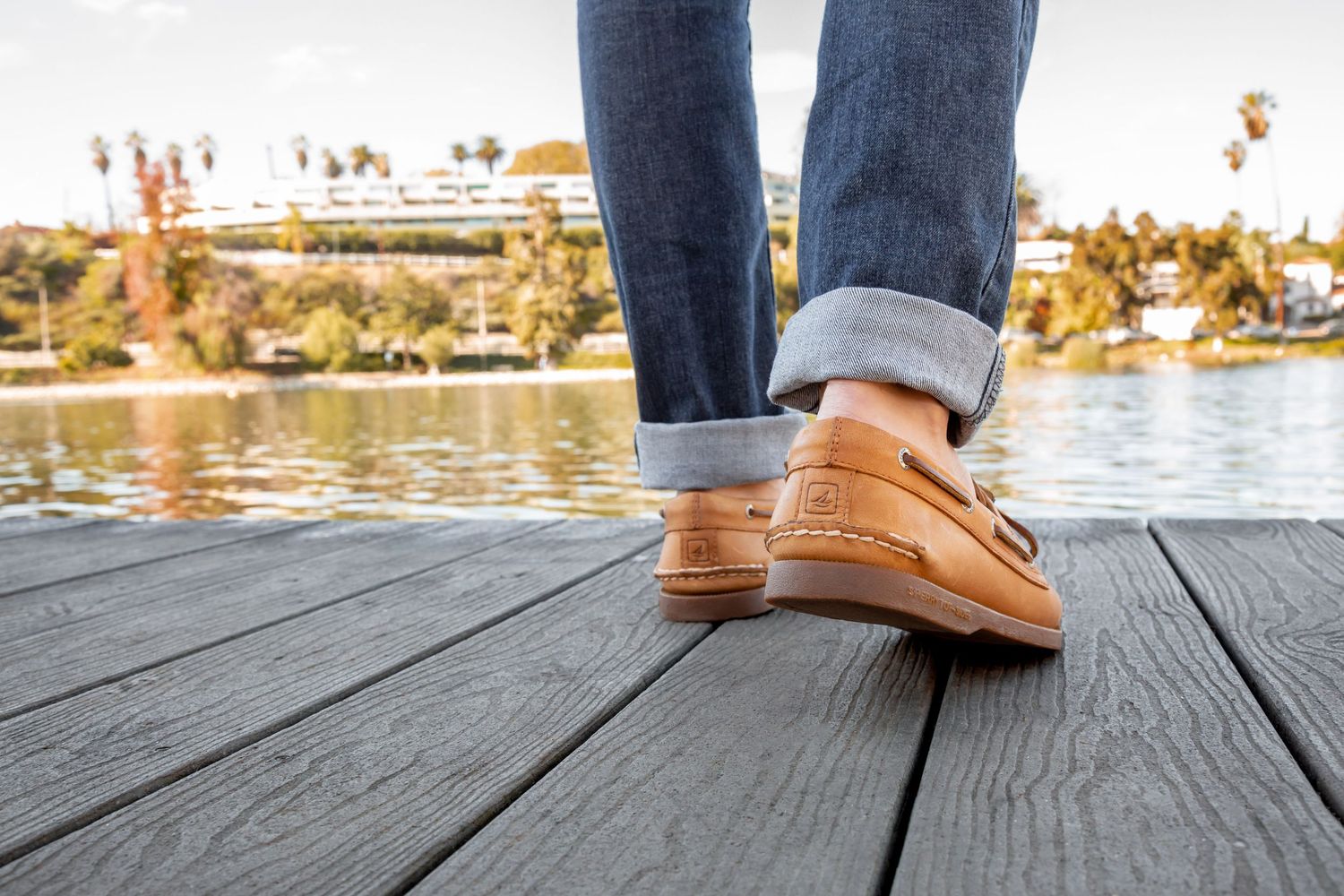 Brown leather boat shoes step across weathered wooden dock planks beside a tranquil lake on a sunny day.