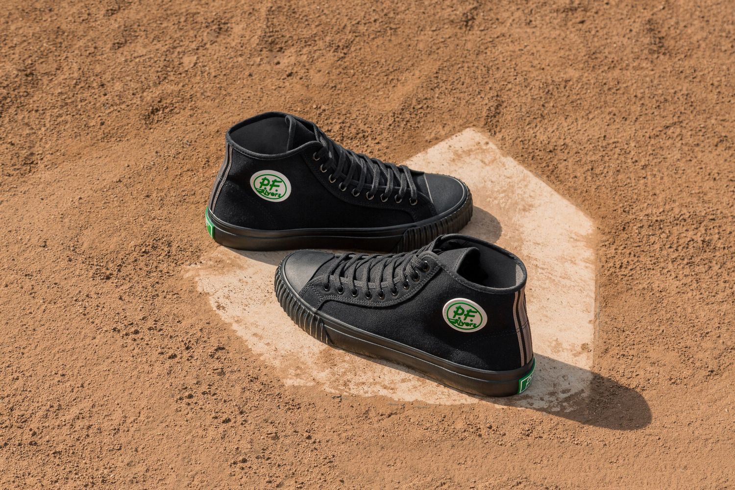 A pair of black high-top PF Flyers sneakers with green logos casting shadows on sandy ground.