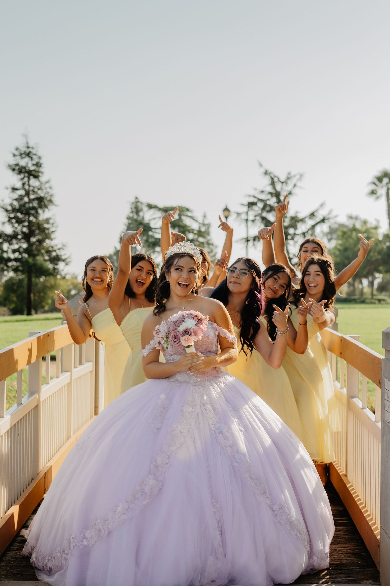 A bridal party in yellow dresses poses joyfully with the bride in a white ballgown on a wooden walkway.
