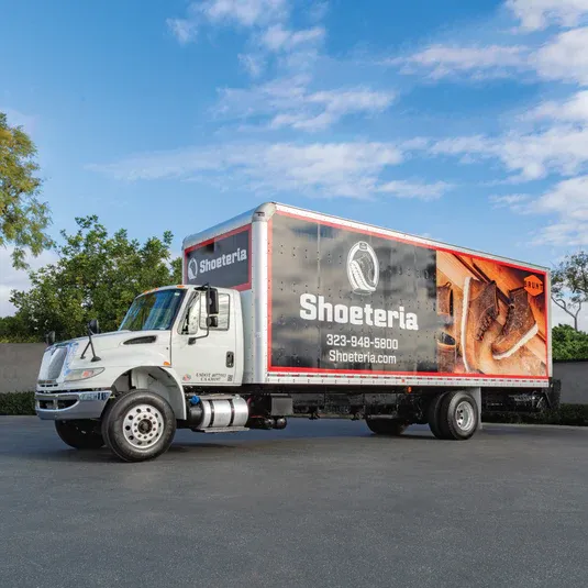 White delivery truck with Shosteria restaurant branding and food imagery parked under blue sky with trees in background.