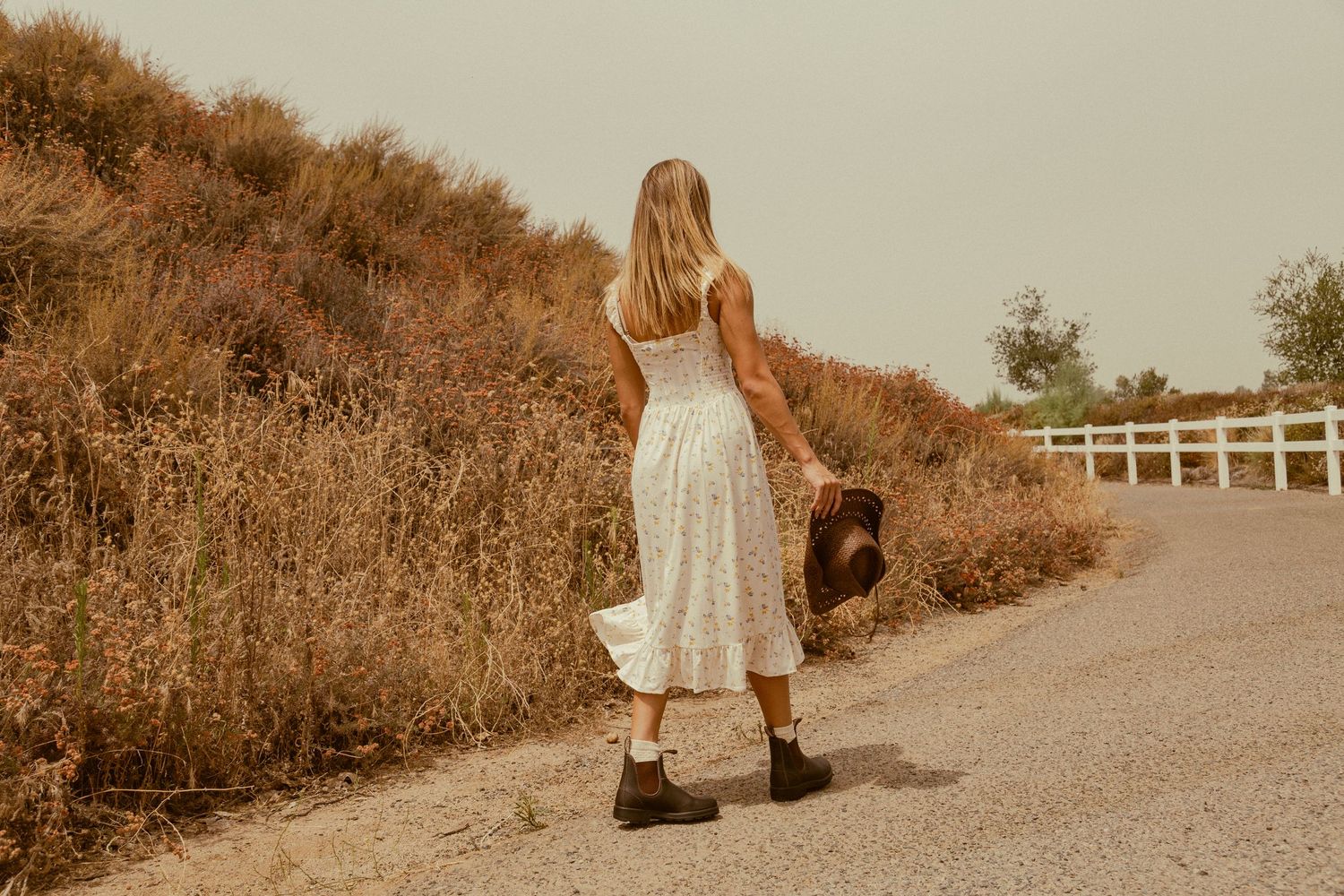 A person in a white dress walks along a sepia-toned dirt path through dried grass and brush.
