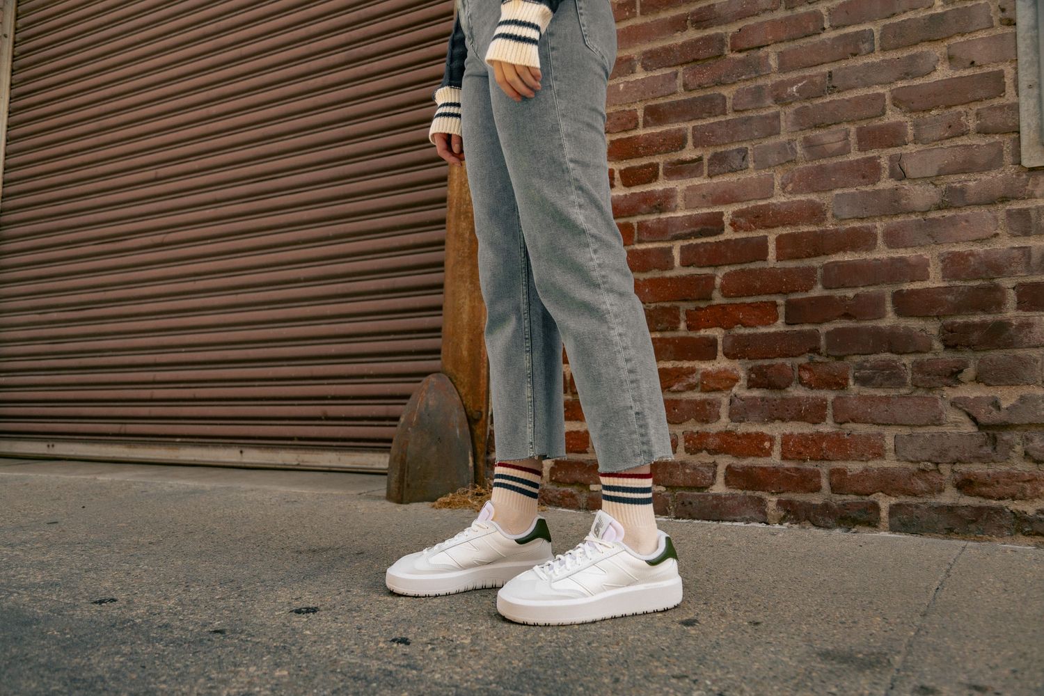 A person wearing jeans and white sneakers stands against a brick wall background.