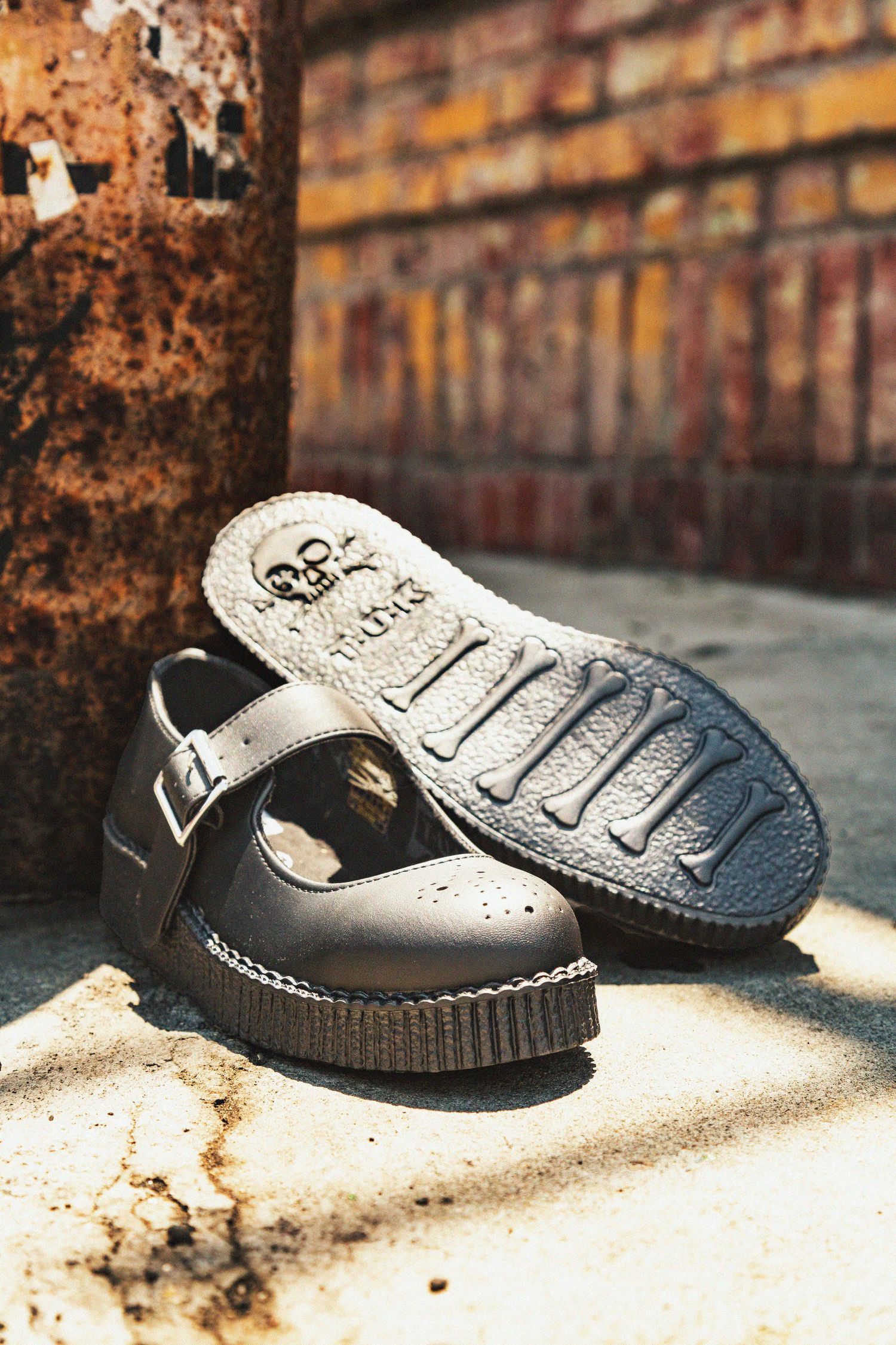 Black leather slip-on shoes with white stitching details sit on a sandy surface against a rustic brick wall background.