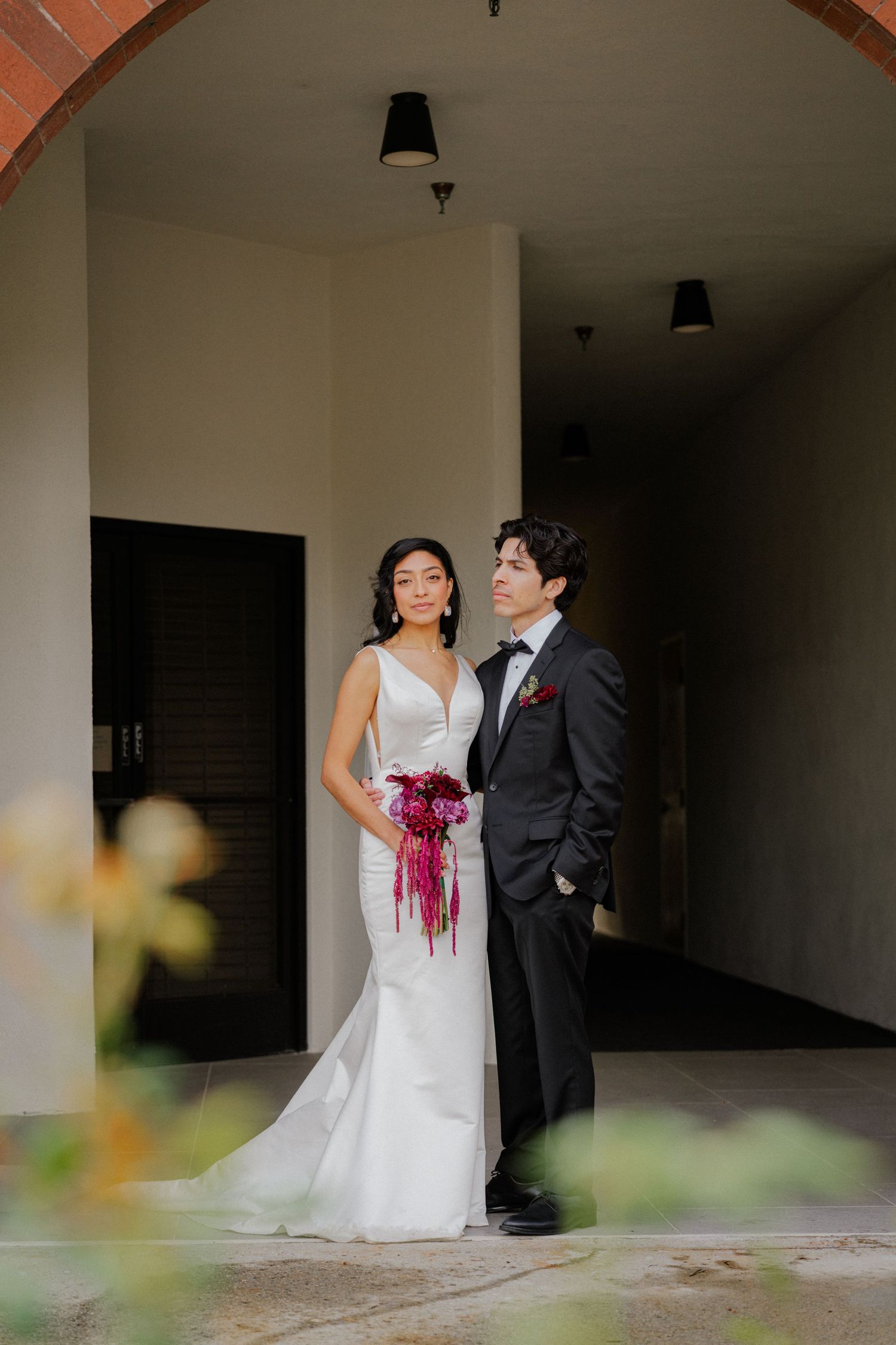 A couple in wedding attire stands together in a hallway with white walls and dramatic lighting.