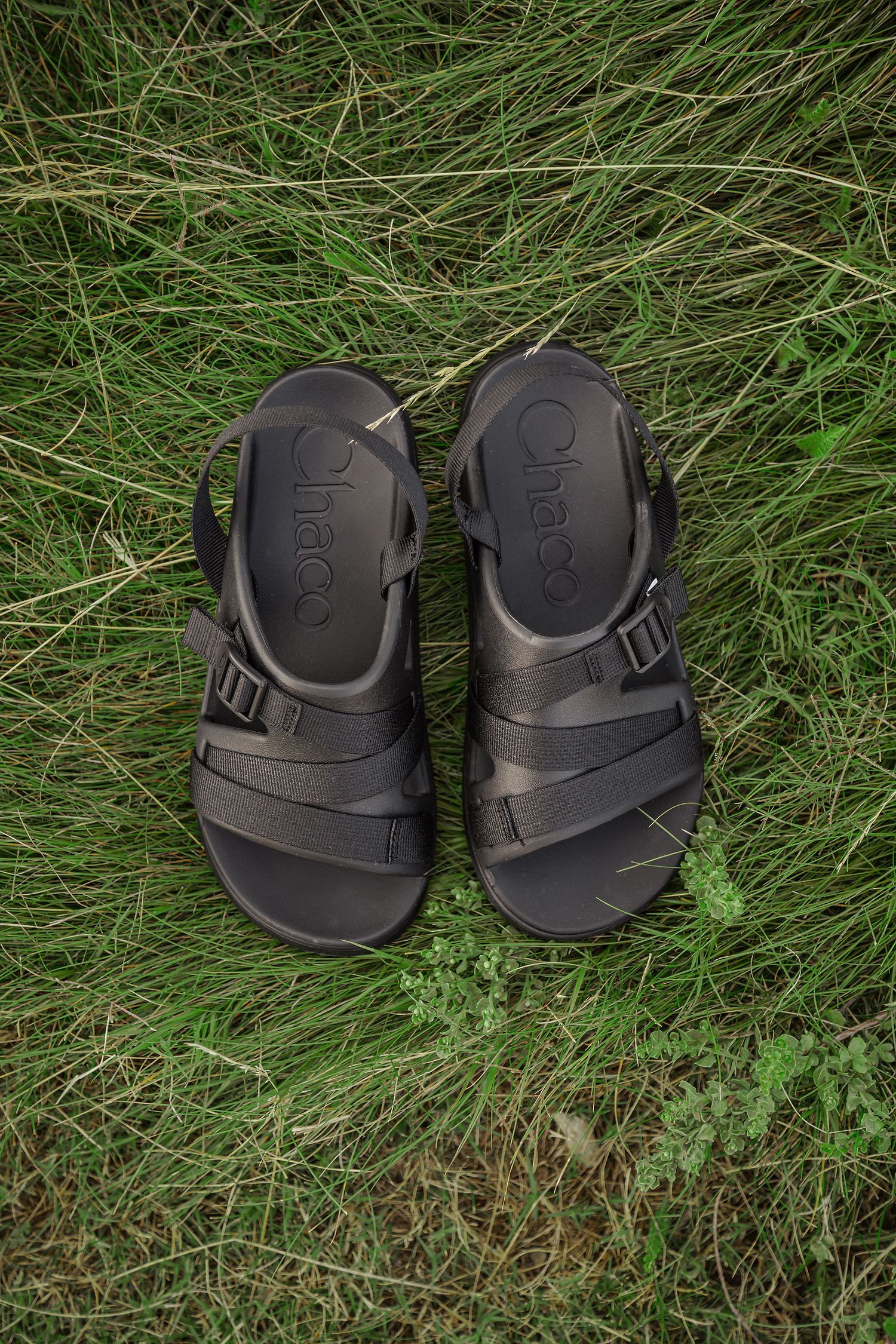 A pair of black plastic sandals with adjustable straps sitting on green grass.