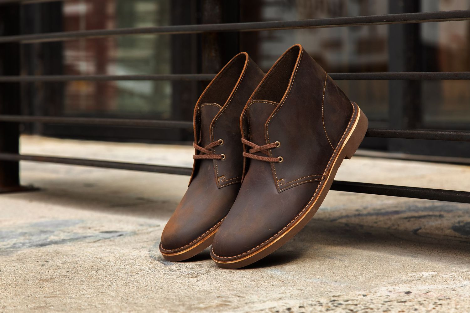 A pair of brown leather chukka boots with contrast stitching sits on a concrete surface near a window.