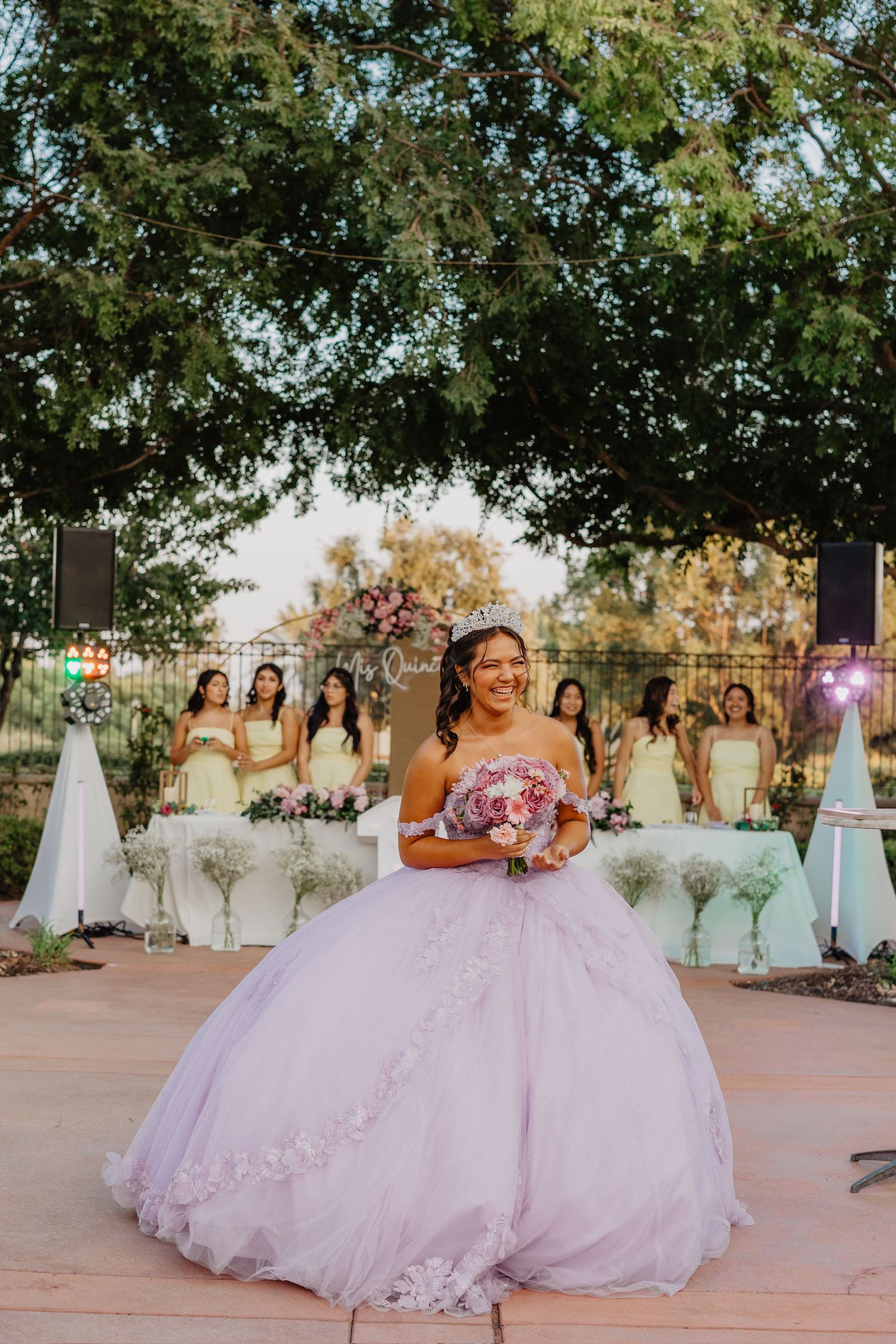 A group of bridesmaids in white dresses stand behind a bride in a pink princess ballgown at an outdoor celebration.