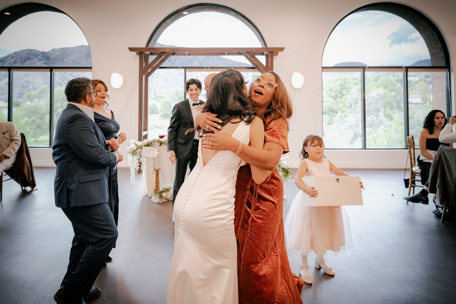 Wedding guests embrace and dance together during a celebration in a mountain venue with arched windows.