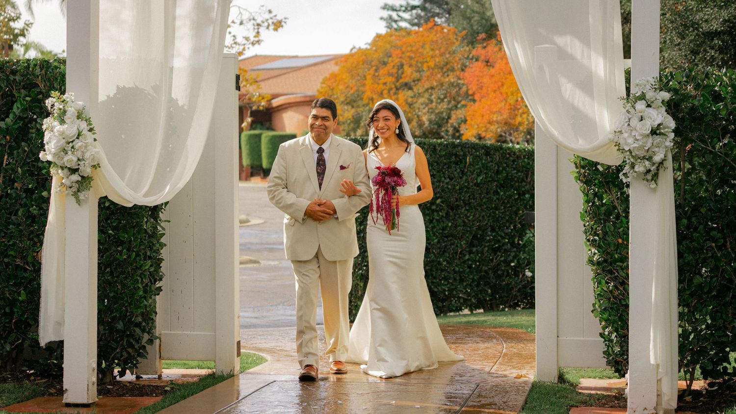 A panoramic view of an elegant white wedding ceremony setup with draped fabric and autumn foliage in the background.