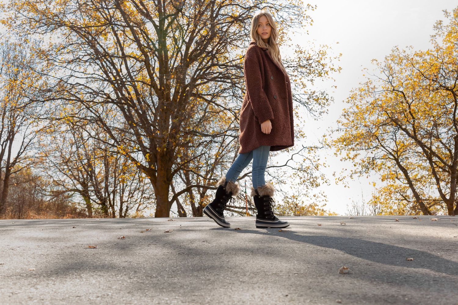 A person in a brown coat and jeans walks on a paved path surrounded by autumn trees with golden leaves.