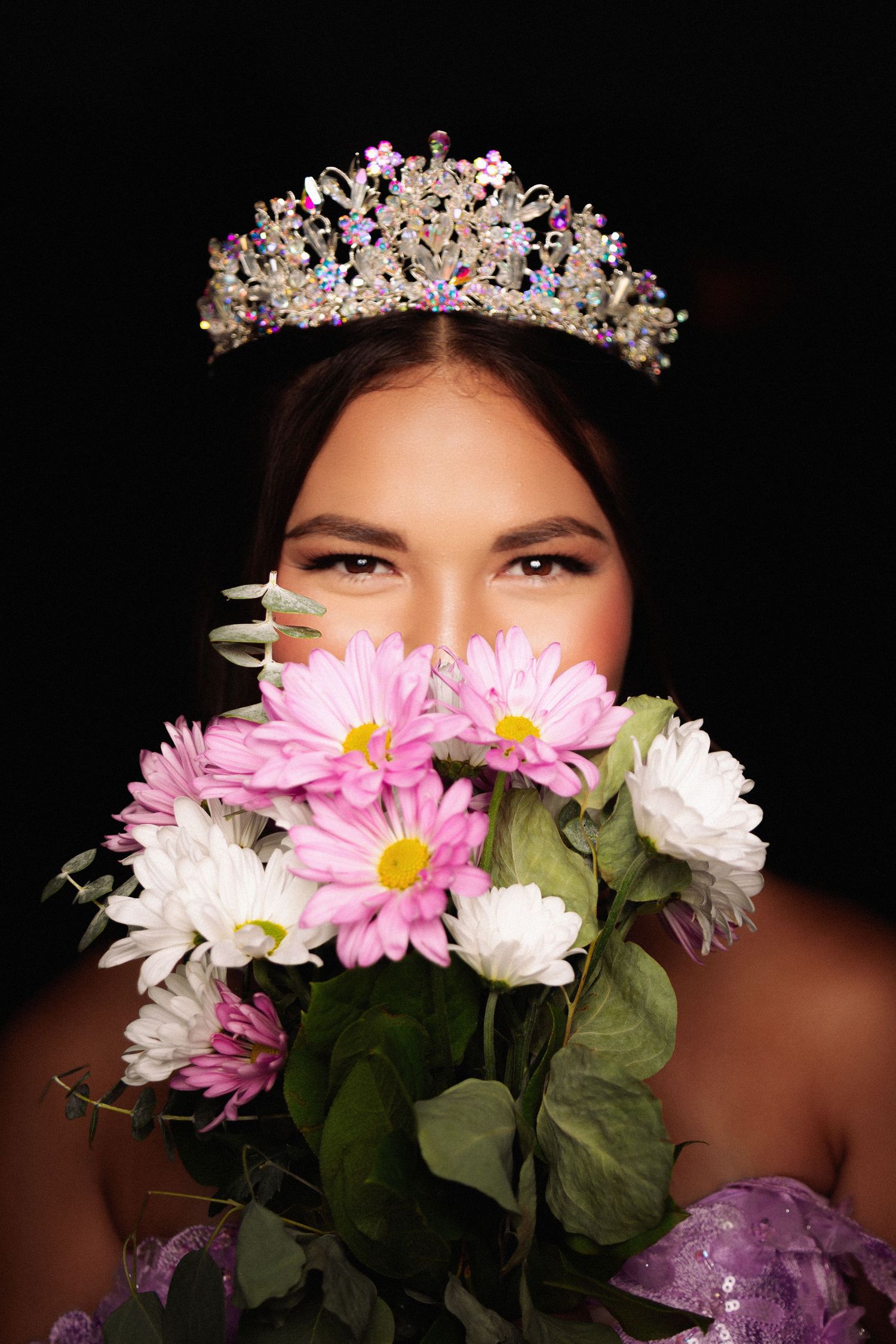 A sparkling tiara crowns a portrait with pink daisies and white flowers against a dark background.