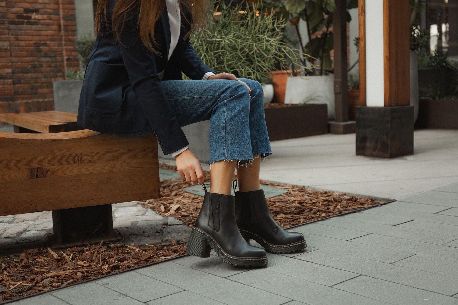 Legs wearing brown leather boots and blue jeans walking along a brick sidewalk in an urban setting.