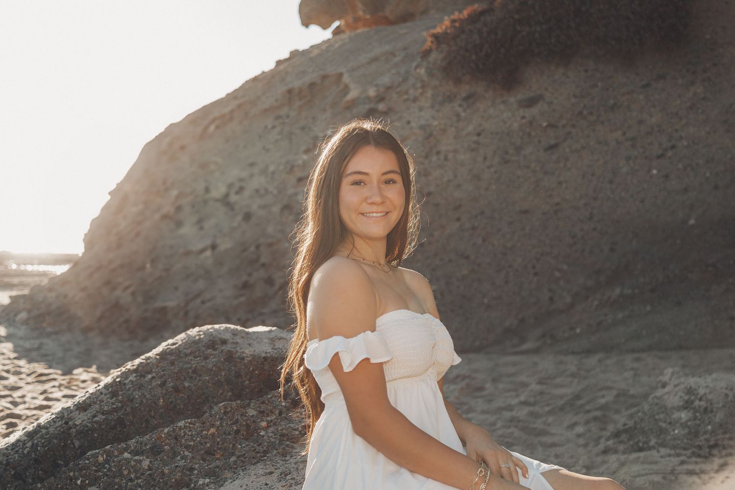 A person in a white dress poses along a rocky coastal cliff during golden hour sunset.