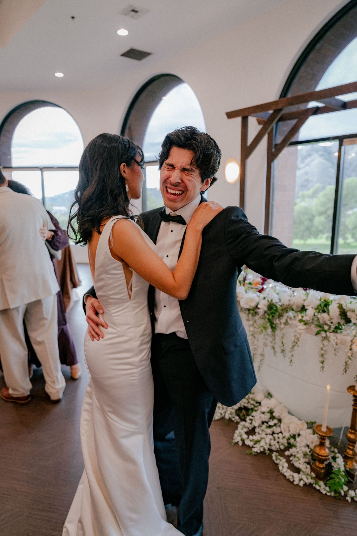 A joyful couple shares a dance moment at their wedding reception in an elegant venue with arched windows and floral decor.