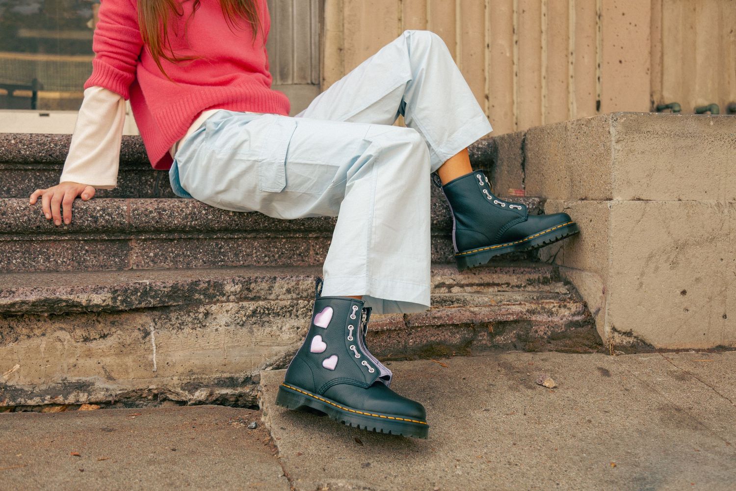 A person in a pink sweater and white pants sits on concrete steps while wearing black chunky loafers.