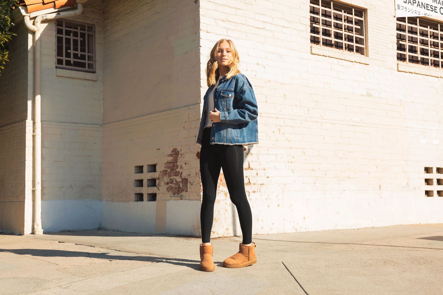 Person in denim jacket and black pants stands in front of white building exterior in natural lighting.