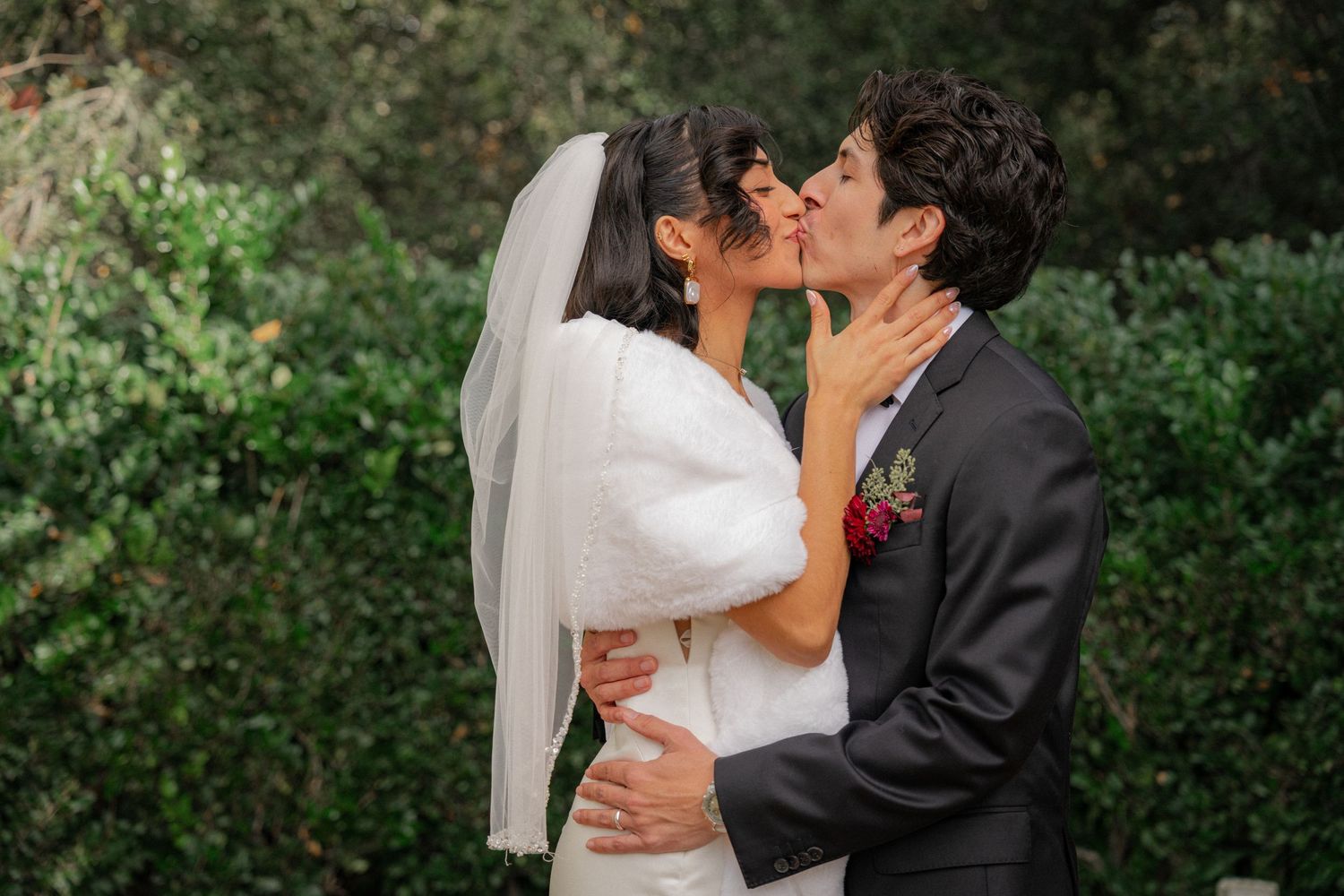 A romantic wedding couple shares a kiss outdoors with greenery in the background.