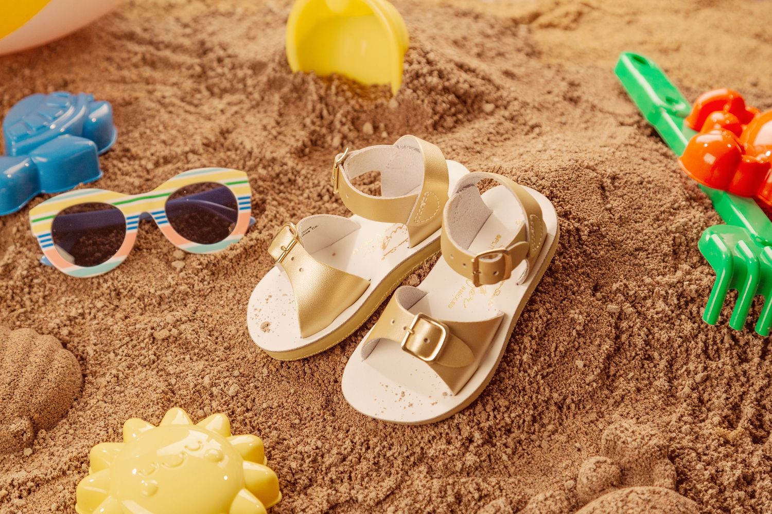 Gold sandals and sunglasses on beach sand with colorful plastic toys scattered around.