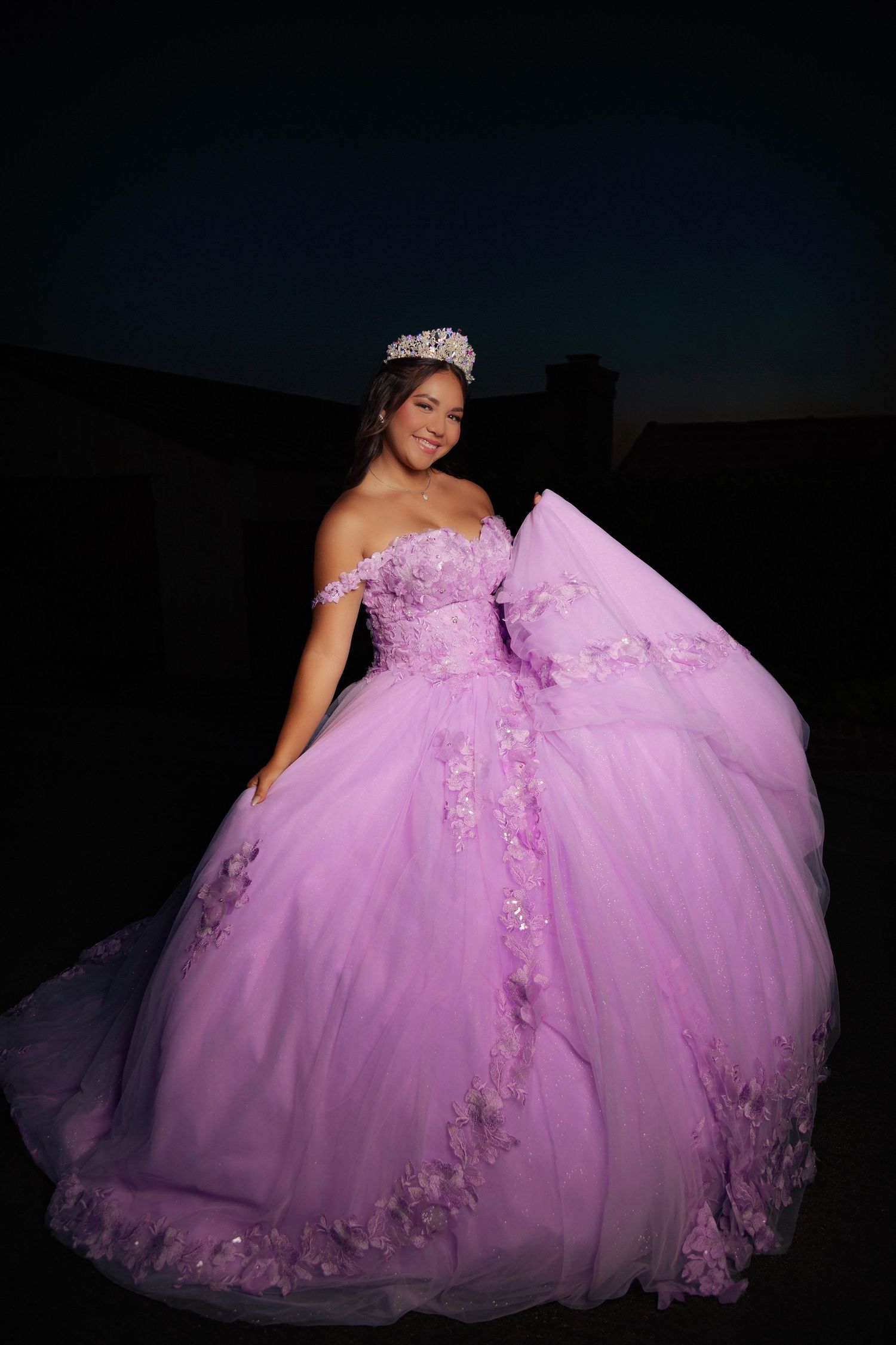 A person in a lavender quinceañera ball gown with off-shoulder design and floral appliques poses against a dark background.