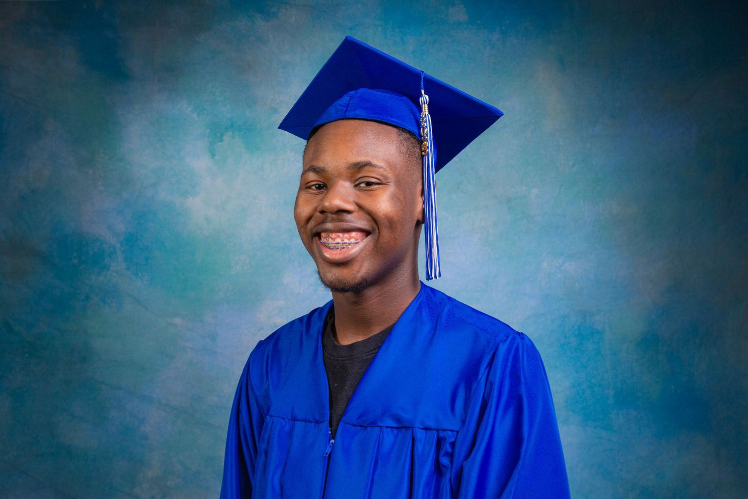 Graduate in bright blue cap and gown smiles warmly against mottled blue studio backdrop.