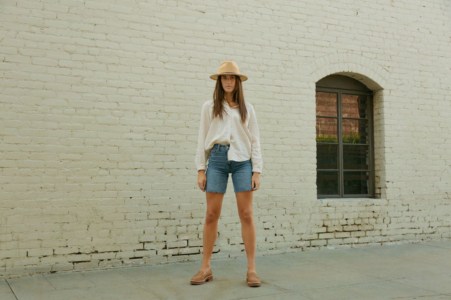 Person in denim shorts and white top stands against a white brick wall in an urban setting.